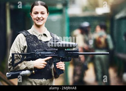 Paintball, felice e ritratto di donna con pistola in uniforme di sicurezza per il gioco di tiro all'aperto con sorriso. Ragazza del torneo eccitata nella protezione del giubbotto Foto Stock