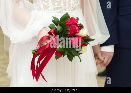 Un bouquet luminoso con rose rosse nelle mani della sposa sullo sfondo di un abito bianco. Foto Stock