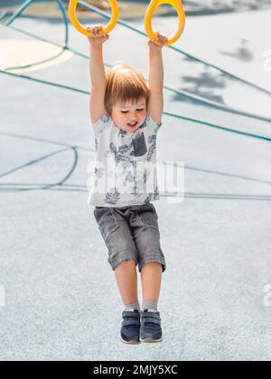 Il ragazzino si sta allenando in campo sportivo pubblico con pavimento blu. Il capretto sta facendo le esercitazioni sugli anelli di sport. Attività all'aperto. Foto Stock