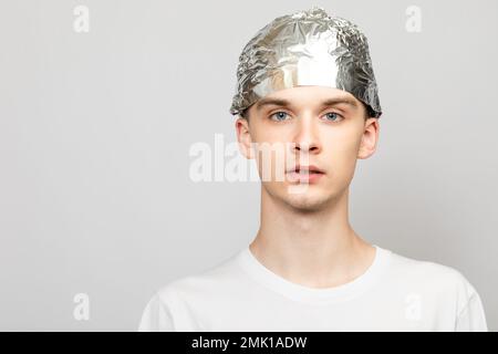 Ritratto di un giovane uomo che indossa un cappello in lamina di stagno. Teorie della cospirazione e concetto di paranoya. Studio girato su sfondo grigio Foto Stock