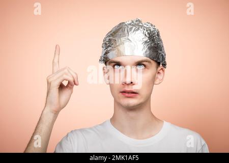 Ritratto di giovane ansioso che indossa un cappello di stagnola puntando il dito e guardando verso l'alto. Teorie della cospirazione e concetto di paranoya. Studio shot sul salmone Foto Stock