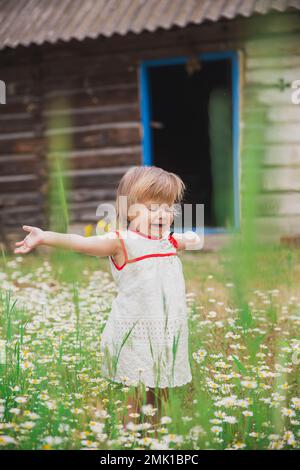 bambino di charme in un vestito ricamato gode di fiori vicino a una capanna di legno Foto Stock