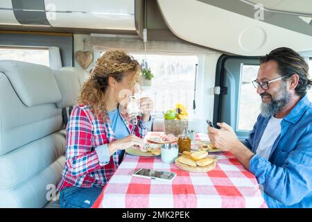 Giovane uomo maturo e donna che si divertono all'interno di un moderno camper mangiando insieme e parlando. Coppia felice nell'attività di viaggio del veicolo. Utilizzando il telefono Foto Stock