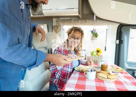 Gentile marito che serve cibo a sua moglie all'interno di una casa camper. Donna sorridente e mangia con il ragazzo. Una coppia felice si diverte a viaggiare al coperto Foto Stock