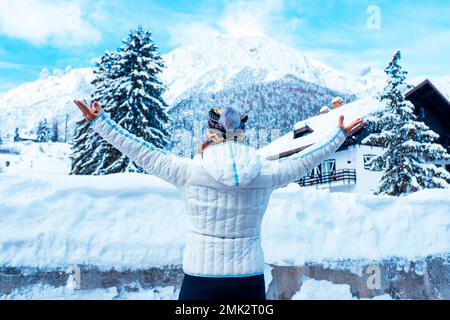 Vista posteriore di una donna turistica che gode di inverno paesaggio di neve montagne e case che si stendono le braccia aperte e ammirando la bellezza. Una donna w Foto Stock