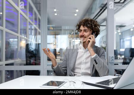 Riuscito uomo d'affari riuscito che comunica allegramente nell'ufficio sul telefono, finanziere ispanico che lavora al posto di lavoro usando il laptop, uomo nel vestito di affari al lavoro felice con il risultato. Foto Stock