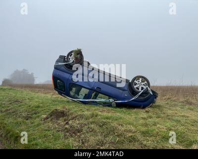 Un'auto hatchback Suzuki rovesciata blu che riposa sul suo tetto dopo essersi schiantata da una strada sull'erba Foto Stock