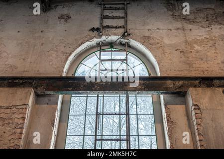 Alta finestra porticato in una vecchia fabbrica. Trave a i in acciaio nella parete in mattoni e scala in metallo fissata alla parete. Paesaggio invernale fuori dalla finestra. Arrugginito, Foto Stock