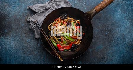 Piatto asiatico mescolare le tagliatelle di udon fritte con verdure e funghi in una teglia nera rustica wok con bacchette di legno su fondo rustico di cemento blu scuro Foto Stock