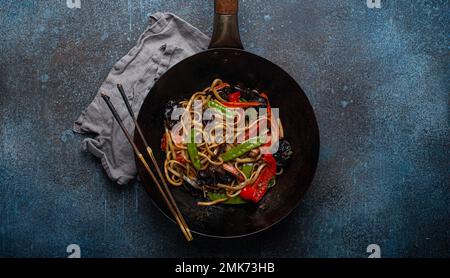 Piatto asiatico mescolare le tagliatelle di udon fritte con verdure e funghi in una teglia nera rustica wok con bacchette di legno su fondo rustico di cemento blu scuro Foto Stock