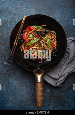 Piatto asiatico mescolare le tagliatelle di udon fritte con verdure e funghi in una teglia nera rustica wok con bacchette di legno su fondo rustico di cemento blu scuro Foto Stock