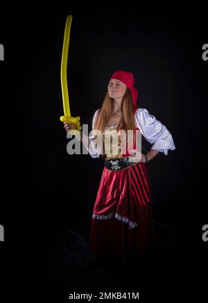 donna con capelli lunghi vestita da pirata in tuta rossa e con un pallone giallo a forma di spada nella fotografia a mano di eventi di festa Foto Stock