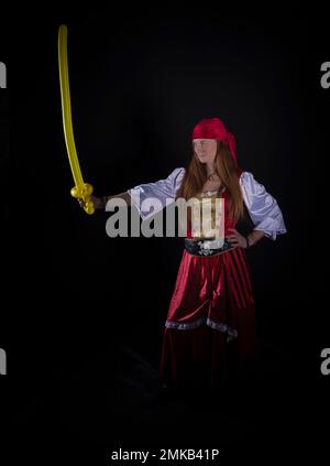 donna con capelli lunghi vestita da pirata in tuta rossa e con un pallone giallo a forma di spada nella fotografia a mano di eventi di festa Foto Stock