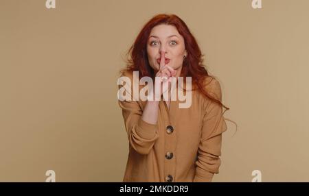 Shh Be Silenzio per favore. Ritratto di donna millenaria di 20 anni preme indice dito a labbra rende silenzio gesto segno non dice segreto. Giovane bella ragazza rossa che posa su sfondo beige studio Foto Stock