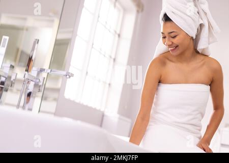 Donna biraciale sorridente che indossa un asciugamano in bagno, che prepara il bagno, con spazio copia. Concetto di relax, cura di sé, bellezza, tempo libero e stile di vita. Foto Stock
