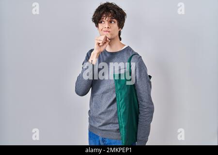 Giovane uomo che indossa una borsa riutilizzabile faccia seria pensando alla domanda con mano sul mento, pensieroso di confondere l'idea Foto Stock