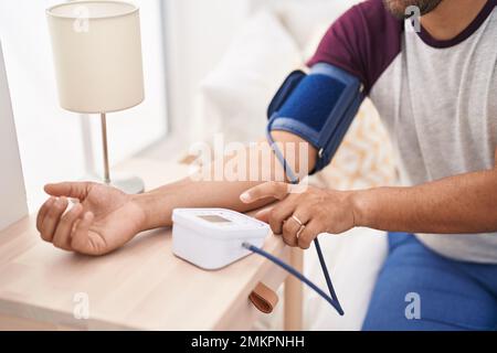 Giovane uomo latino che misura l'impulso usando il tensiometro in camera da letto Foto Stock
