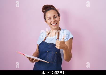 Bruna donna che indossa cameriera professionale grembiule tenere appunti sorridendo felice e positivo, pollice su facendo eccellente e segno di approvazione Foto Stock