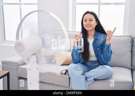 Giovane donna asiatica bere un bicchiere d'acqua godendo aria dal ventilatore sorridendo felice indicando con mano e dito al lato Foto Stock