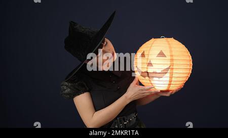 Giovane donna cinese che indossa il costume della strega che tiene la lampada della zucca di Halloween sopra lo sfondo nero isolato Foto Stock