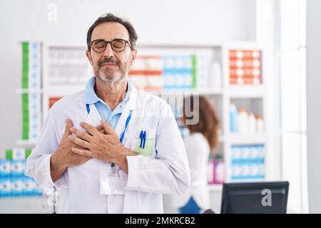 Uomo ispanico di mezza età che lavora in farmacia sorridendo con le mani sul petto, gli occhi chiusi con gesto grato sul viso. concetto di salute. Foto Stock