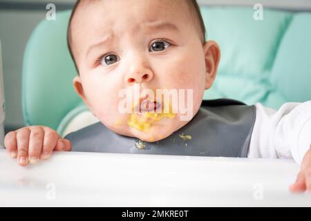 Un bambino sorpreso sporco macchiato in purea di verdure al tavolo di alimentazione. Introduzione di alimenti complementari, non piace il gusto, nuovo cibo. Primo piano Foto Stock