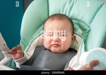 Il bambino è cattivo al tavolo sul seggiolone. Il bambino affamato sconvolto, rifiuta di mangiare, introduzione di alimenti complementari Foto Stock