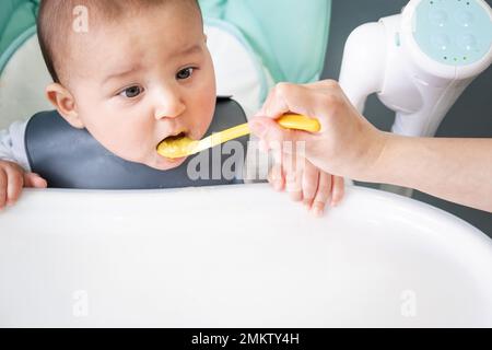 La mamma nutre il bambino con un cucchiaio di purea di verdure al tavolo per l'allattamento dei bambini. Appetito del bambino, nutrizione sana, introduzione di complementare Foto Stock