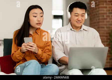Problema coniugale. Donna asiatica interessata che guarda il laptop del marito e usa il cellulare, seduto insieme sul divano Foto Stock