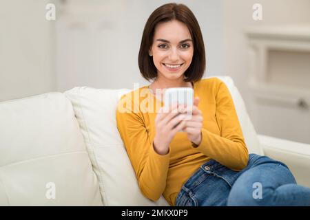 Ragazza simpatica che riposa sul divano a casa, utilizzando lo smartphone Foto Stock