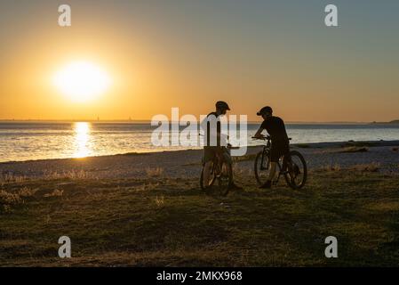 Due ciclisti retroilluminati che parlano al tramonto sulla spiaggia di Sletterhage, Djursland, Jutland, Danimarca Foto Stock