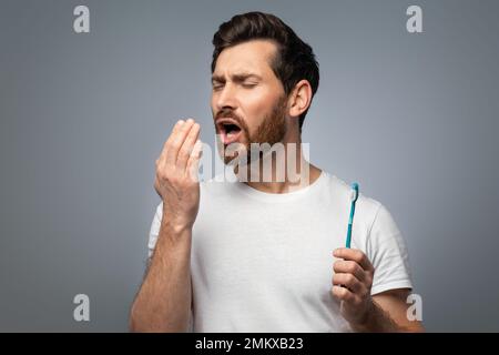 Alito cattivo. Bell'uomo di mezza età che controlla il respiro con la mano, soffiando su di esso, in piedi su sfondo grigio Foto Stock