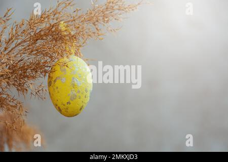 Uovo di Pasqua giallo appeso su ramo di erba secca su fondo grigio. Raggi luminosi di sole. Buona Pasqua. Scheda con spazio di copia per il testo. Foto Stock