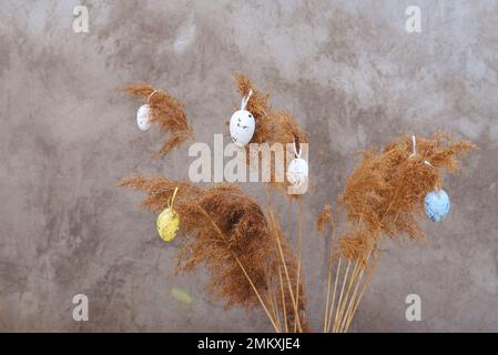 Fuoco selettivo morbido sul bouquet di erba di canna appeso con le uova di Pasqua colorate su fondo grigio. La vista è diritta. Buona Pasqua. Scheda con spazio di copia per il testo. Foto Stock