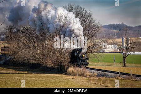 Una vista di un classico treno passeggeri a vapore che si avvicina, viaggiando attraverso la campagna, soffiando fumo e vapore in un giorno d'inverno Foto Stock