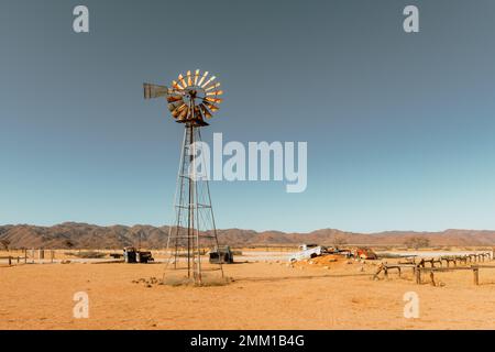 Abbandonato derelitto vecchio cimitero auto e vecchia turbina eolica arrugginita nel deserto sabbioso con un cielo blu sullo sfondo Foto Stock