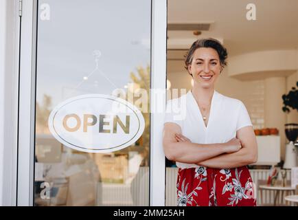 Il proprietario di un caffè di successo sorride alla macchina fotografica mentre si trova accanto a un cartello aperto alla porta del suo ristorante. Donna piccola impresa proprietario welcomin Foto Stock
