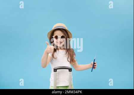 Allegra bambina in t-shirt bianca, cappello di paglia e occhiali da sole, porta la carta d'imbarco, si trova vicino a una valigia, si mostra guardando la macchina fotografica, isolato blu Foto Stock