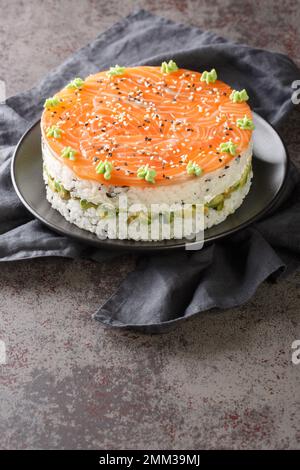 Torta di sushi al salmone con riso, avocado, formaggio cremoso, alghe, semi di sesamo decorati con wasabi primo piano in un piatto sul tavolo. Verticale Foto Stock
