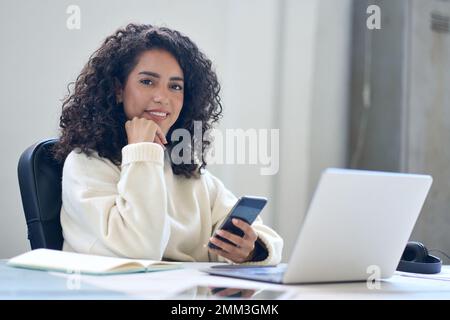 Giovane donna di affari latina felice usando il telefono che lavora in ufficio alla scrivania. Foto Stock