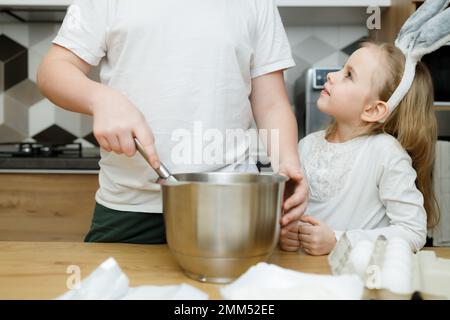 Ragazza piccola in orecchie coniglietto guardando il suo fratello grande mescolarsi, frustando pasta di biscotti in cucina moderna. I fratelli si trovano insieme al tavolo da cucina, mak Foto Stock