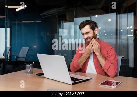 Un giovane uomo, uomo d'affari, freelance, designer siede in ufficio al tavolo. funziona su un computer portatile. Tiene la guancia, sente fastidio in bocca, ha un forte mal di denti. Foto Stock