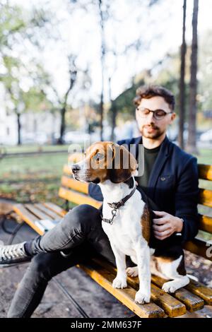 un giovane uomo alla moda siede con il suo cane da aquila su una panca del parco Foto Stock