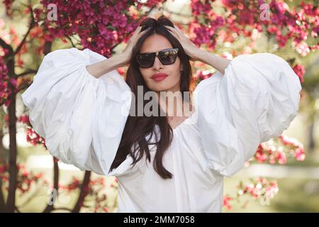 Splendida bella donna brunetta con capelli lunghi in camicia di cotone bianco con maniche larghe lanterne flounce su sfondo primavera sakura fiori. Modello Foto Stock