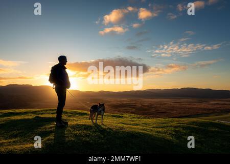 Persona con un cane sulla cima del forte Dunadd Hill al tramonto, vicino a Lochgilphead, Argyll, Scozia, Regno Unito Foto Stock