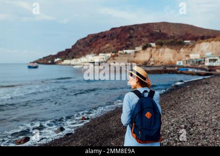 Donna viaggiatore con zaino a piedi sulla spiaggia sull'isola di Santorini, Grecia. Turistico ammirando il mare e il paesaggio montano Foto Stock