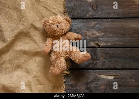 un vecchio orso bruno peloso giace da solo su un pavimento in legno su un panno marrone, un giocattolo per bambini sul pavimento Foto Stock