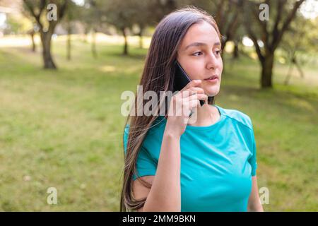 Ragazza studentesca adolescente che indossa una t-shirt acquamarina, facendo una telefonata alla sua famiglia nel parco. Retroilluminazione del sole Foto Stock