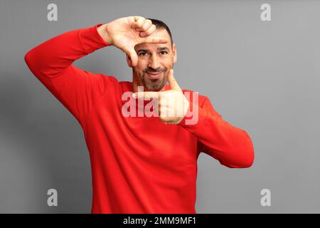 Bel maschio felice bearded gli occhi e guarda attraverso una cornice formata dalle sue mani. Un uomo attraente fa una cornice con le sue dita, isolato Foto Stock