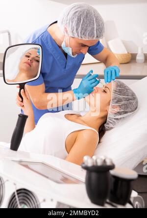 Cliente femminile che guarda in specchio durante la mesoterapia del viso Foto Stock